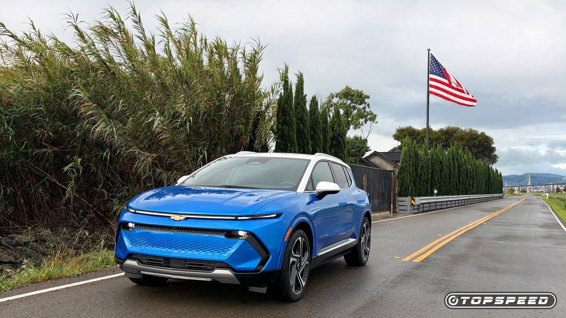 Blue and white Chevrolet Equinox EV parked on a wet road beside tall plants and an American flag.