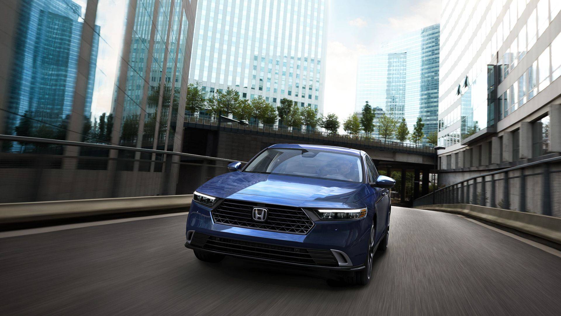 Blue Honda Accord sedan driving through a city street lined with modern glass buildings.