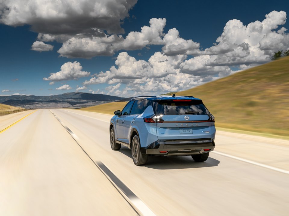 Blue 2027 Nissan Rogue Hybrid driving on a highway through a mountainous landscape