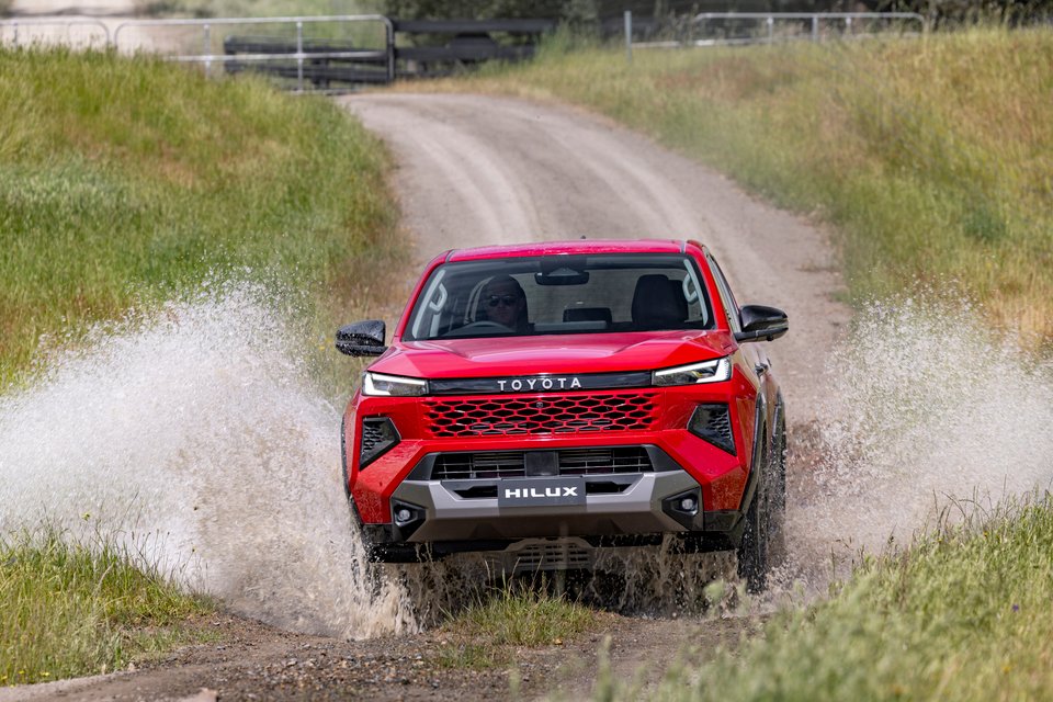 Red Toyota HiLux driving through a shallow water crossing on a gravel track.