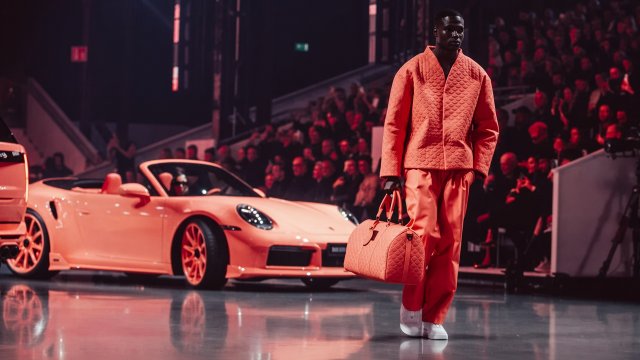 Model in a coral outfit carrying a matching bag beside a BRABUS 900 PEETCH at a runway presentation